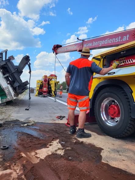 Schwerlast-Bergefahrzeug mit Kran führt eine Lkw-Bergung auf der Autobahn durch, Einsatzkraft in Warnkleidung sichert den Kran, verunfallter Lkw neben der Fahrbahn bei sonnigem Wetter.