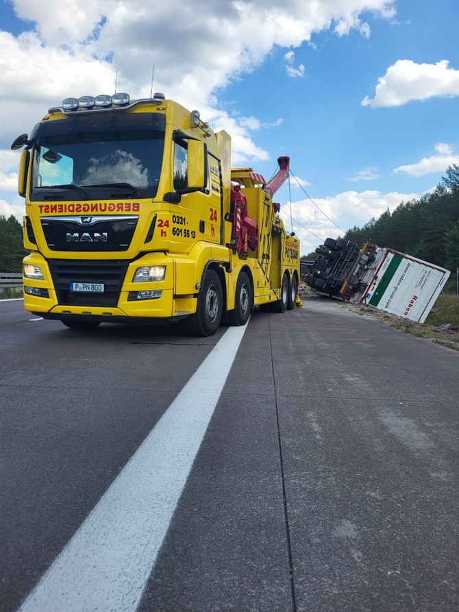 Gelber Schwerlast-Berge-Lkw von MAN steht auf der Autobahn während einer Lkw-Bergung, umgekippter Sattelauflieger liegt im Straßengraben, gesicherte Unfallstelle bei Tageslicht.